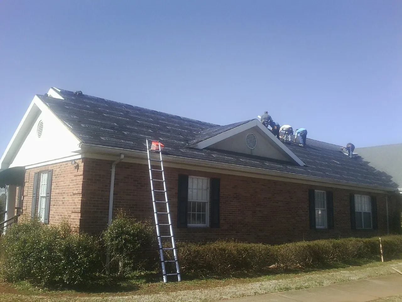 Crew tearing shingles off a brick ranch roof with a ladder set — Emergency Roof Repair work by our crew in Aquebogue