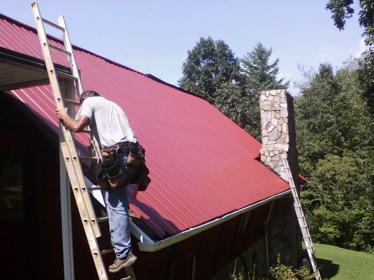 Roofer on a ladder beside a red metal roof and stone chimney — Soffit & Fascia Repair by Roofing Brothers in Aquebogue